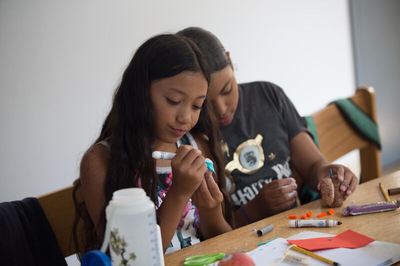 The image shows two people, likely a mother and daughter, focused on crafting at a table. The girl is holding a marker and appears to be working on a small project. Various craft supplies, including markers and small objects, are scattered on the table. The scene suggests a moment of creativity and bonding.